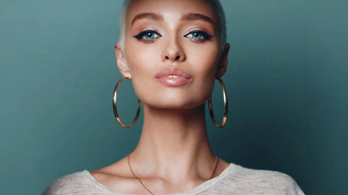 Stilvolle Frau mit platinblondem Haar, die große goldene Creolen und eine zarte Halskette von Anadolu Juweliere trägt und eleganten Schmuck in Düsseldorf präsentiert.