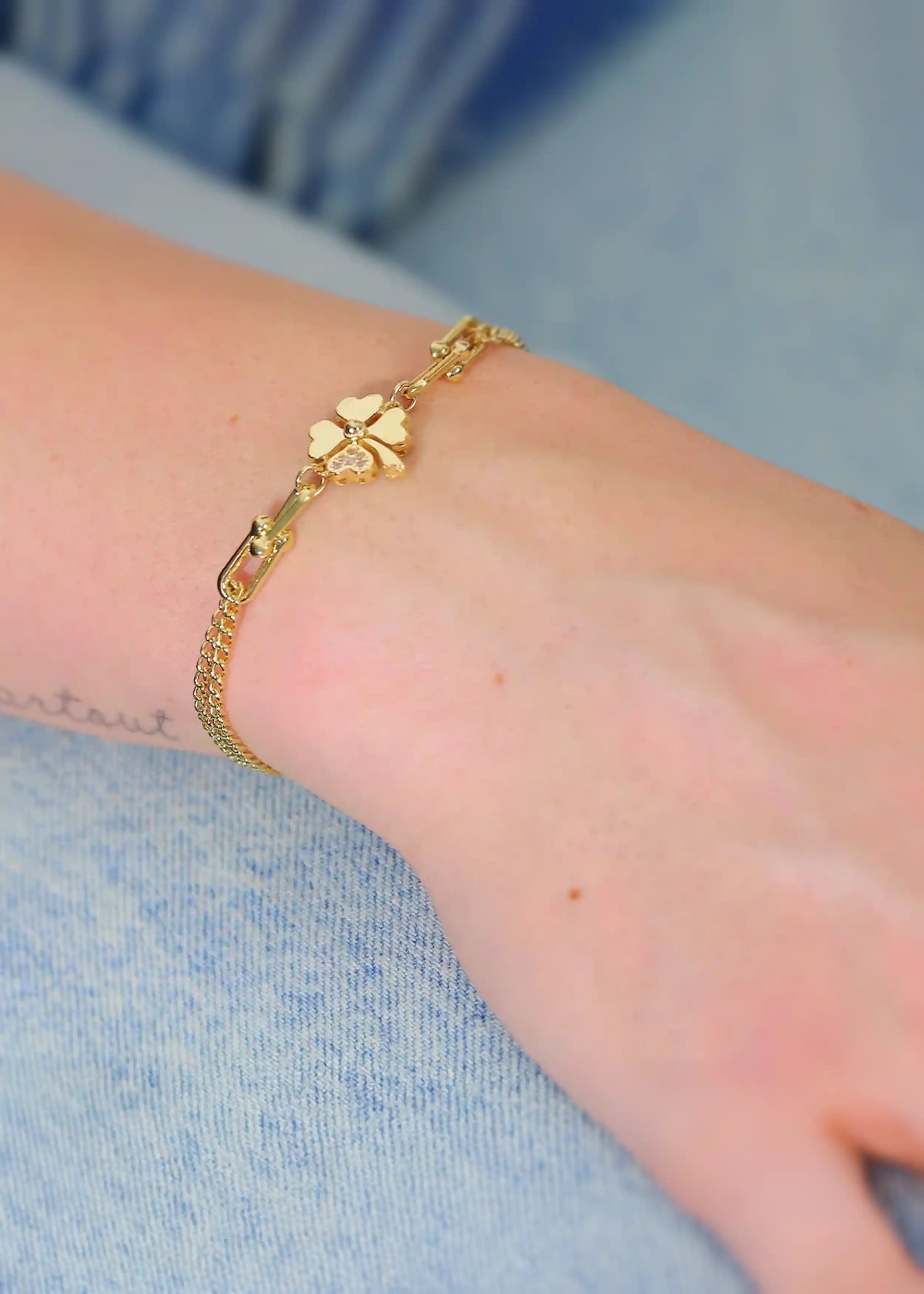 Goldenes Armband mit doppelter Gliederkette und einem vierblättrigen Kleeblatt-Anhänger aus vier Herzchen, besetzt mit funkelnden Steinen, am Handgelenk einer Person. Im Hintergrund ist ein hellblaues Jeanshemd sichtbar.
