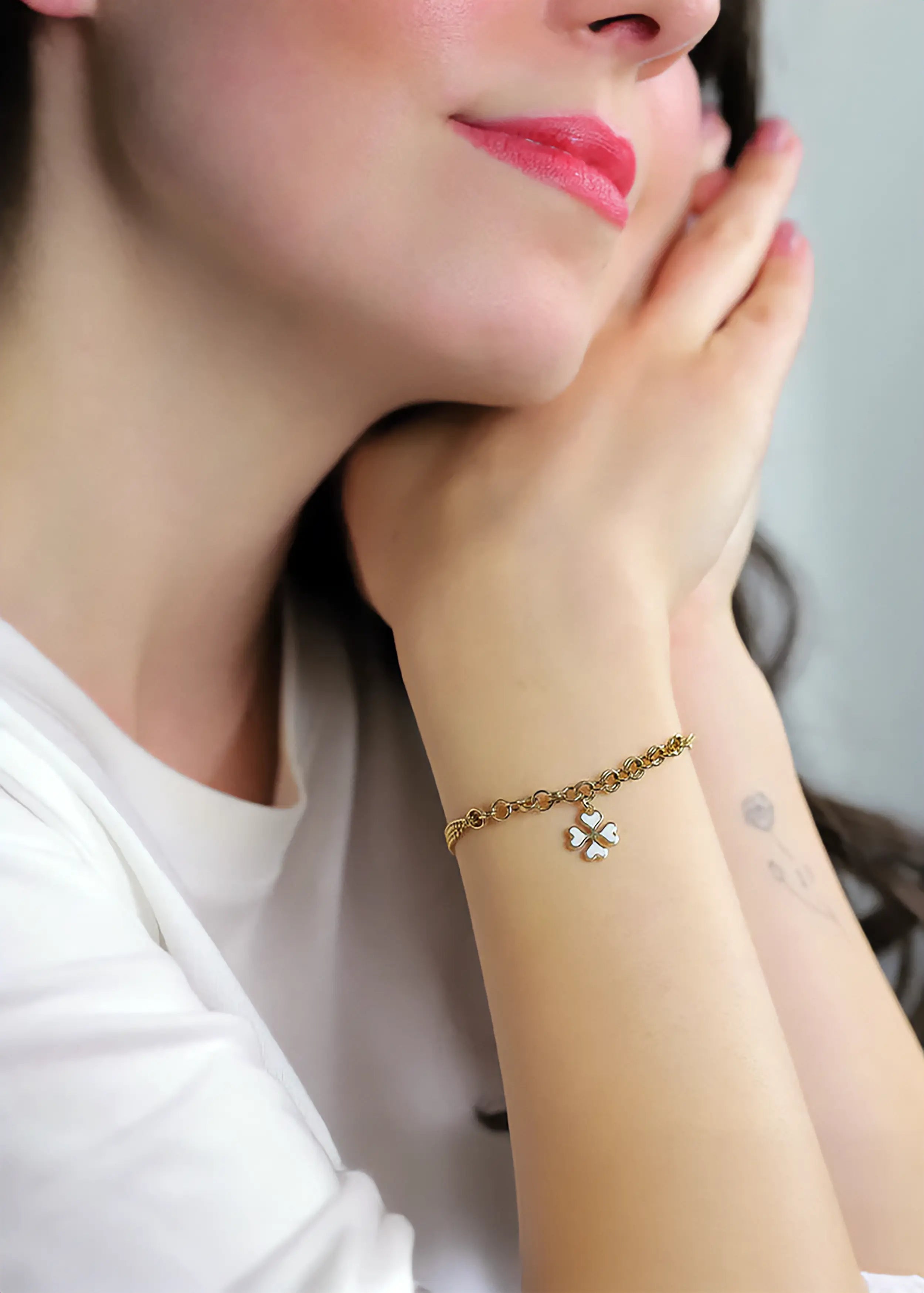 Ein Model trägt Goldenes Armband mit doppelter Gliederkette und einem vierblättrigen Kleeblatt-Anhänger aus weißen Herzchen, auf weißem Hintergrund. Die Kette hat einen Karabinerverschluss und eine Verlängerungskette mit einem runden Anhänger