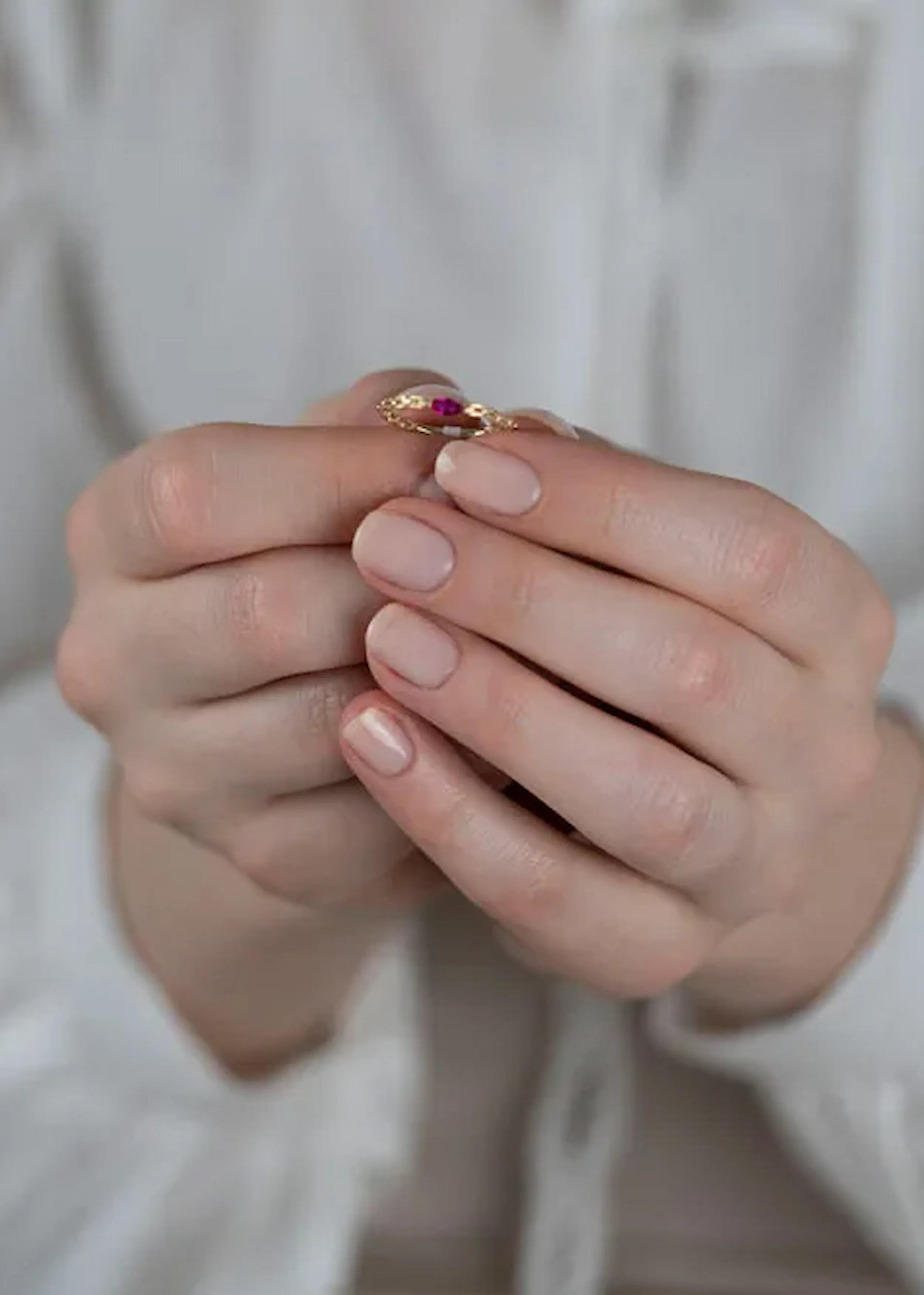 Goldener Ring mit Gliederkette und einem ovalen pink-roten Stein, gehalten in den Händen einer Person mit naturfarbenen Nägeln. Die Person trägt ein weißes Kleidungsstück