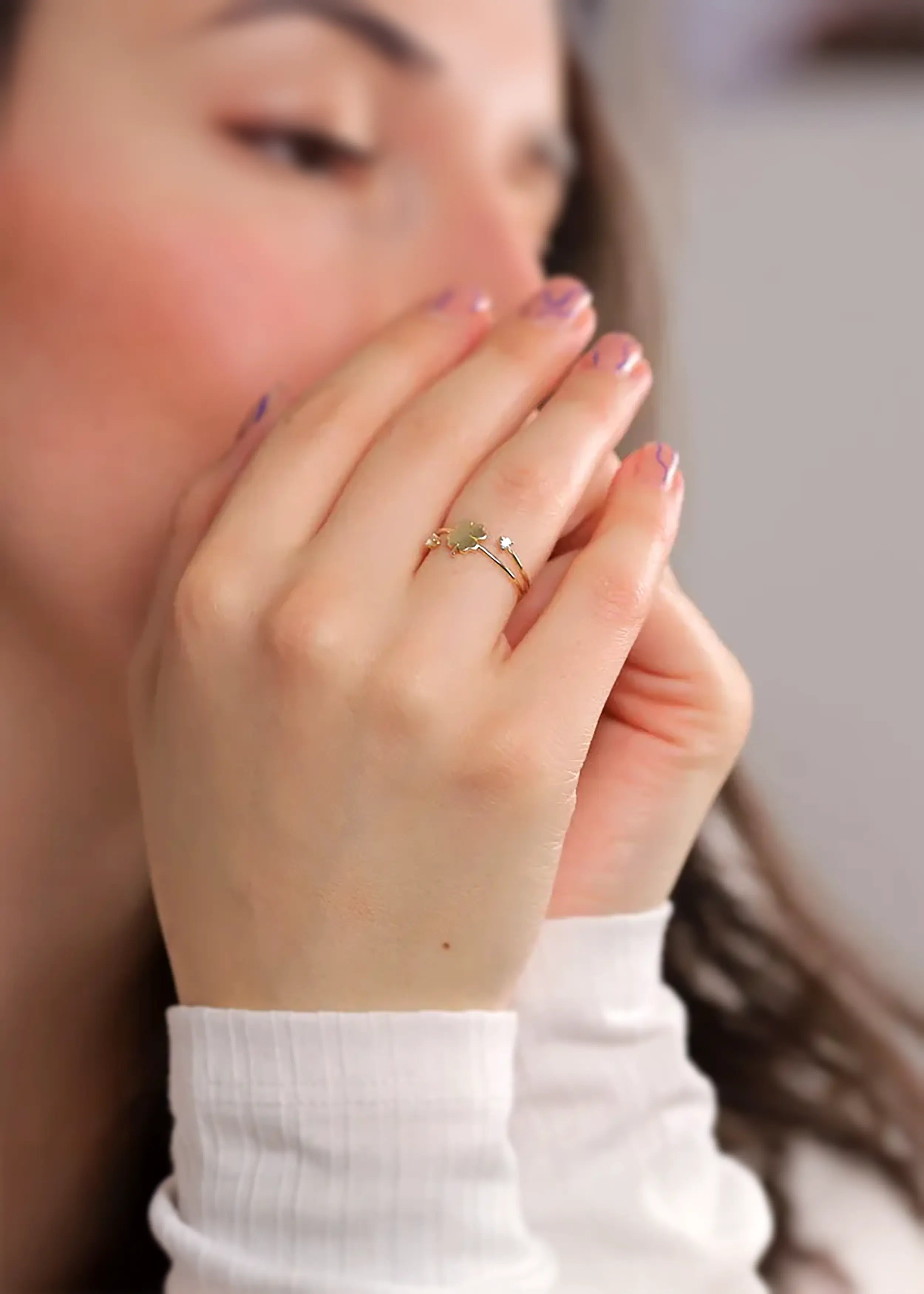 Goldener Ring mit geflochtenem Band und einem vierblättrigen Kleeblatt-Anhänger aus herzförmigen Elementen mit weißem Emaille, am Finger einer Person. Die Person trägt ein weißes Kleidungsstück und hat naturfarbene Nägel.
