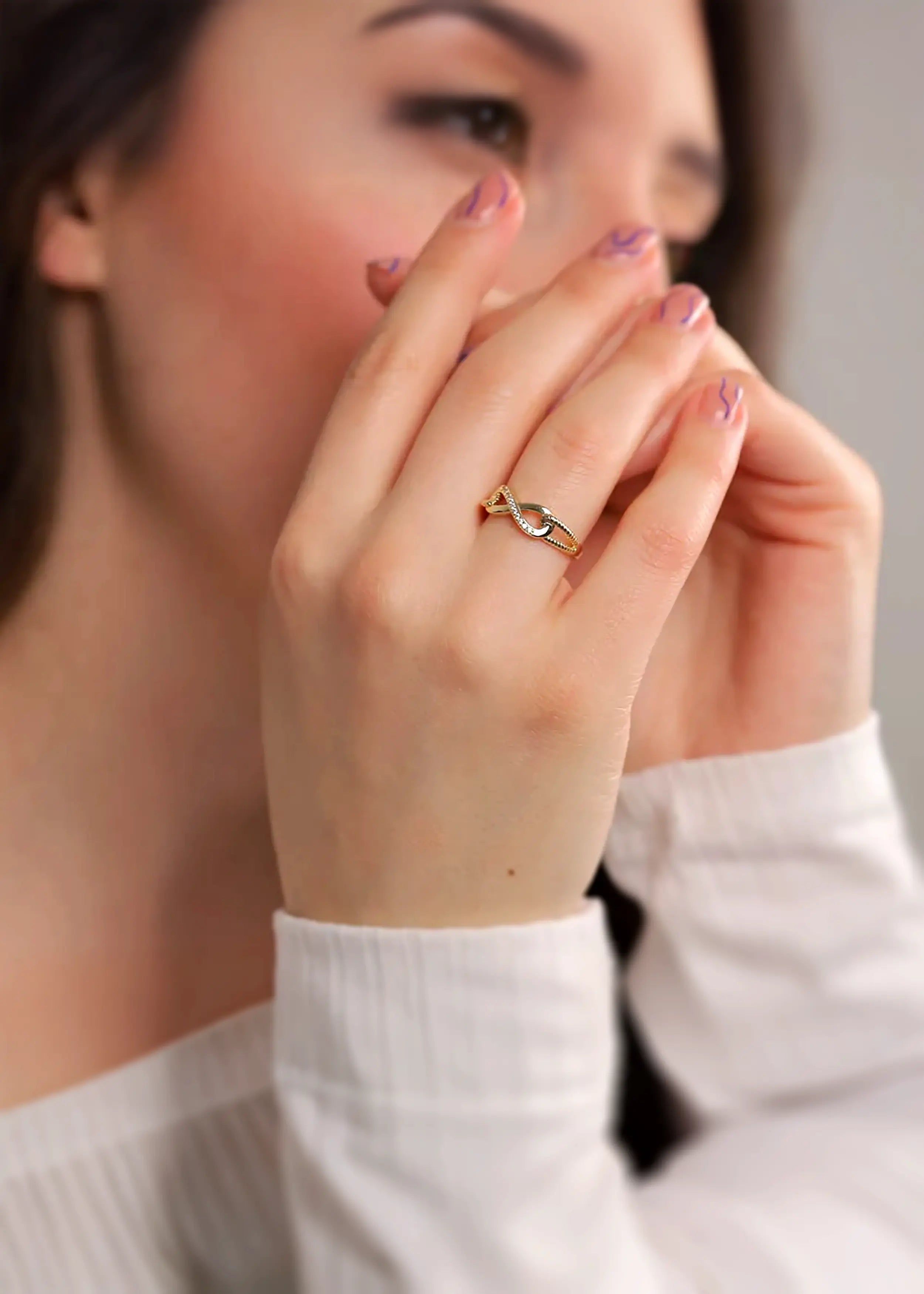 Goldener 14-Karat-Infinity-Ring mit Zirkonia-Steinen, am Finger einer Person. Die Person trägt ein weißes Kleidungsstück und hat naturfarbene Nägel mit blauen Mustern.