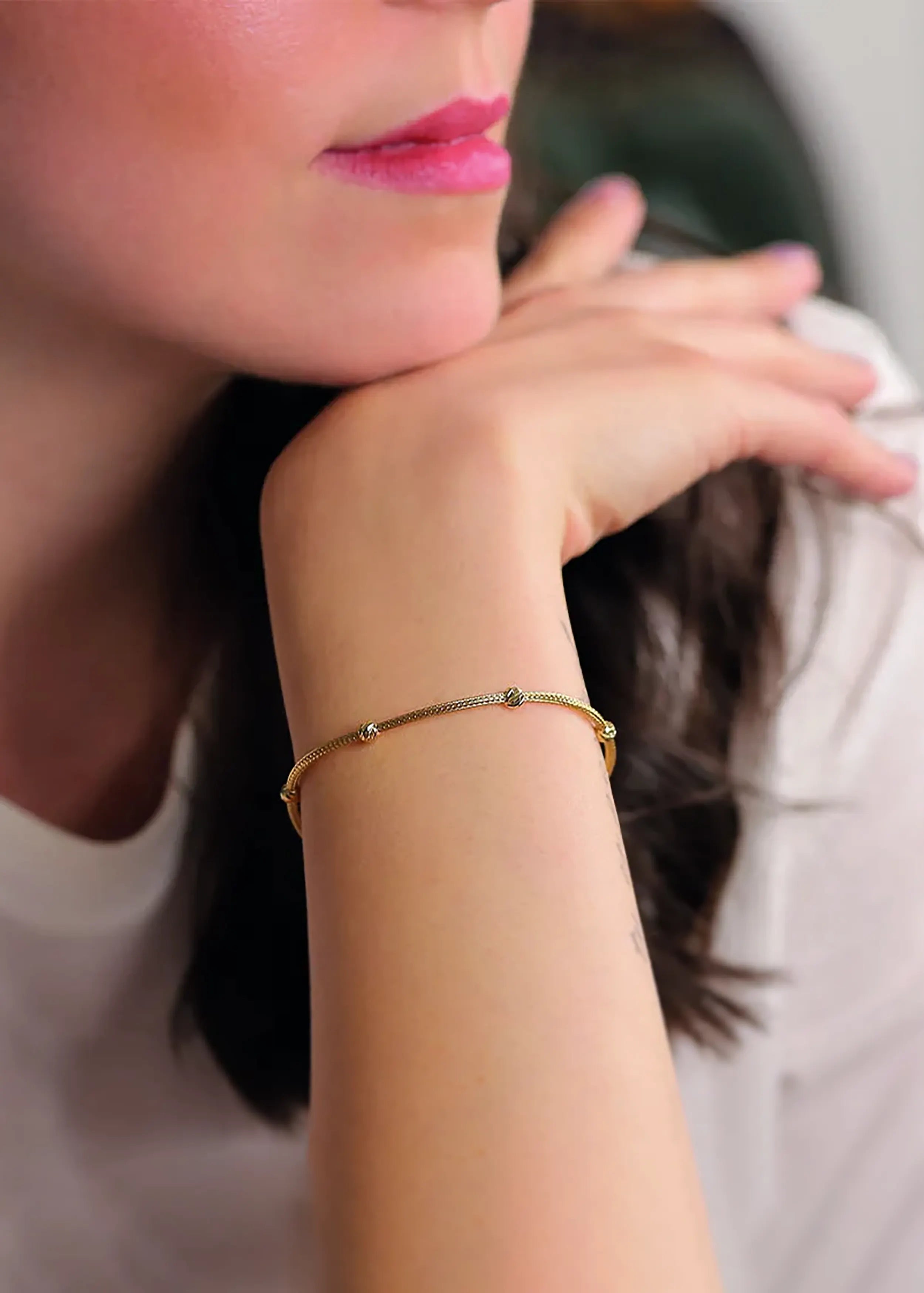 Goldenes Armband mit geflochtener Kette, runden Sech goldene Gugeln und Anhänger, am Handgelenk einer Person. Die Person trägt ein weißes Kleidungsstück und hat pinke Nägel