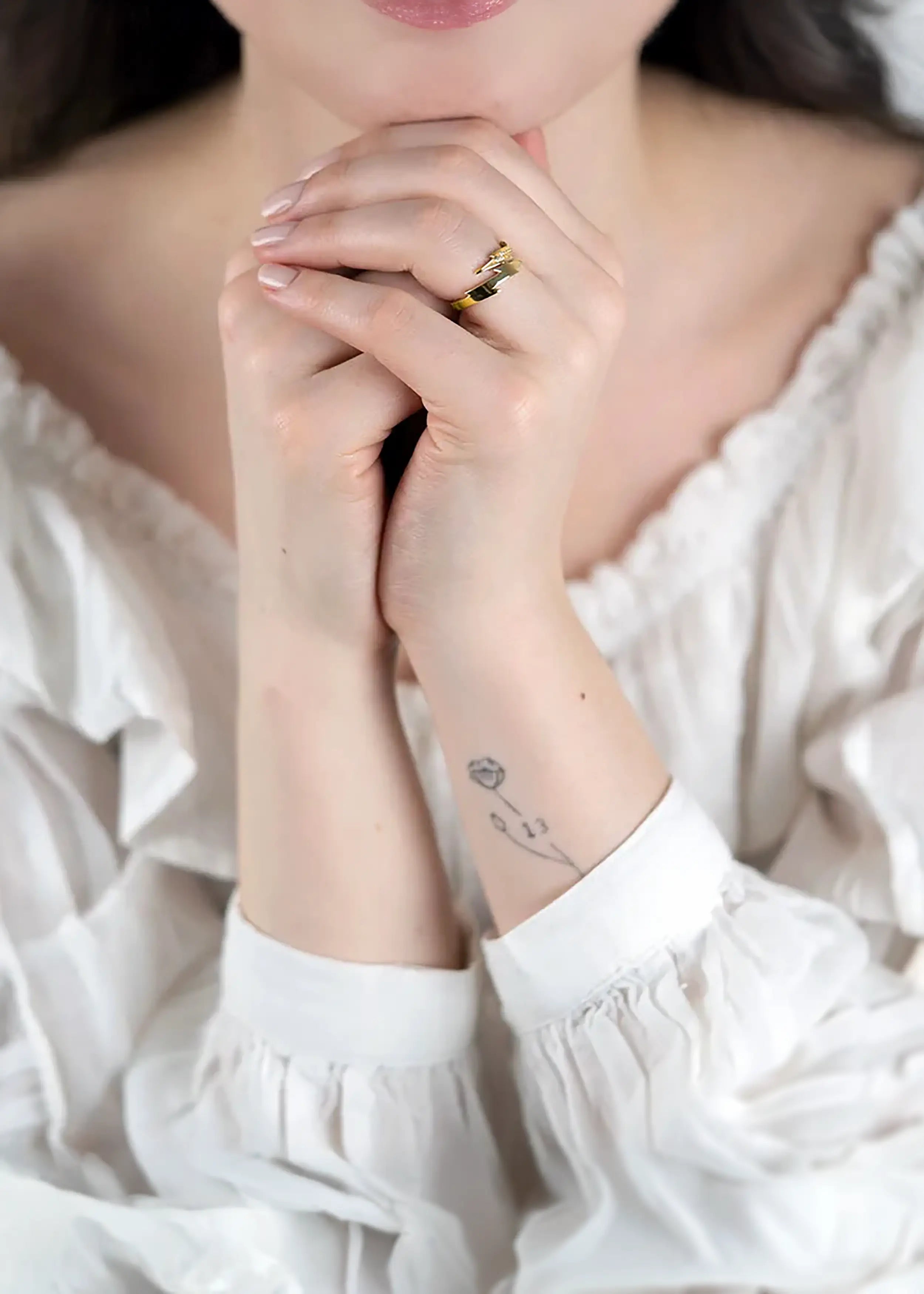 Goldener 14-Karat-Ring mit Blitz-Design und funkelnden Steinen, am Finger einer Person. Die Person trägt ein weißes Kleid mit Rüschen und hat naturfarbene Nägel, mit einem kleinen Tattoo am Handgelenk.