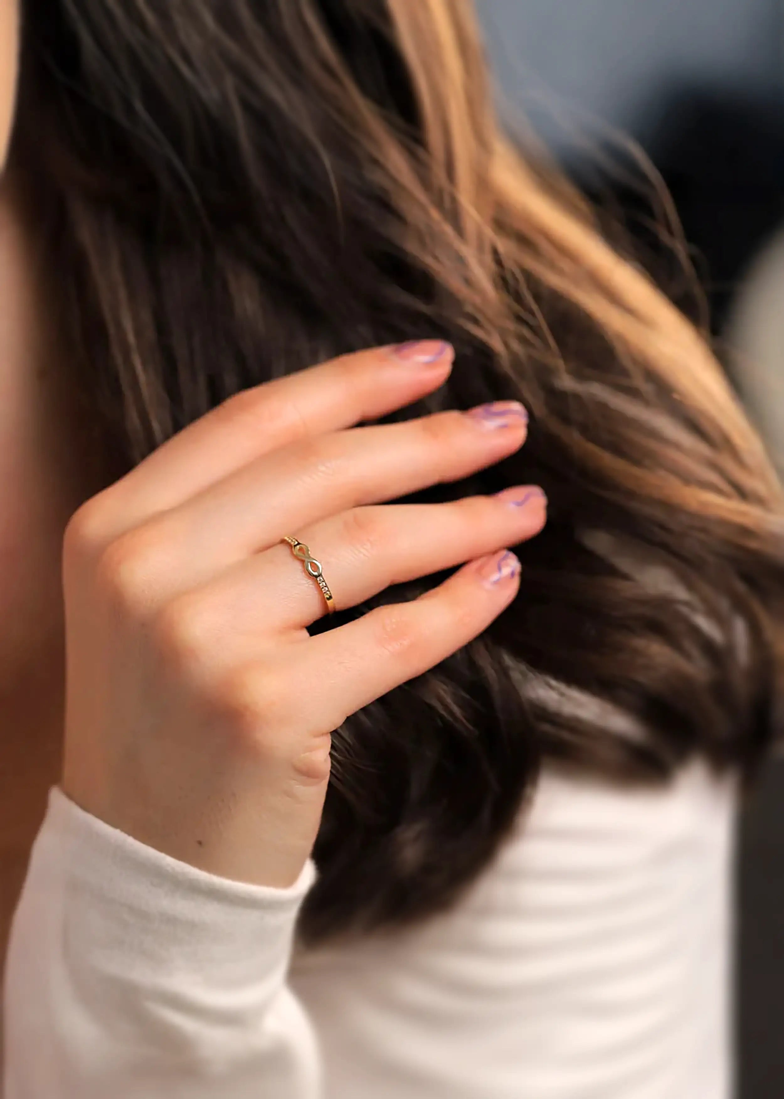 Goldener 14-Karat-Infinity-Ring mit Zirkonia-Steinen, am Finger einer Person. Die Person trägt ein Kleid mit floralem Muster und einen goldenen Armreif, mit naturfarbenen Nägeln.