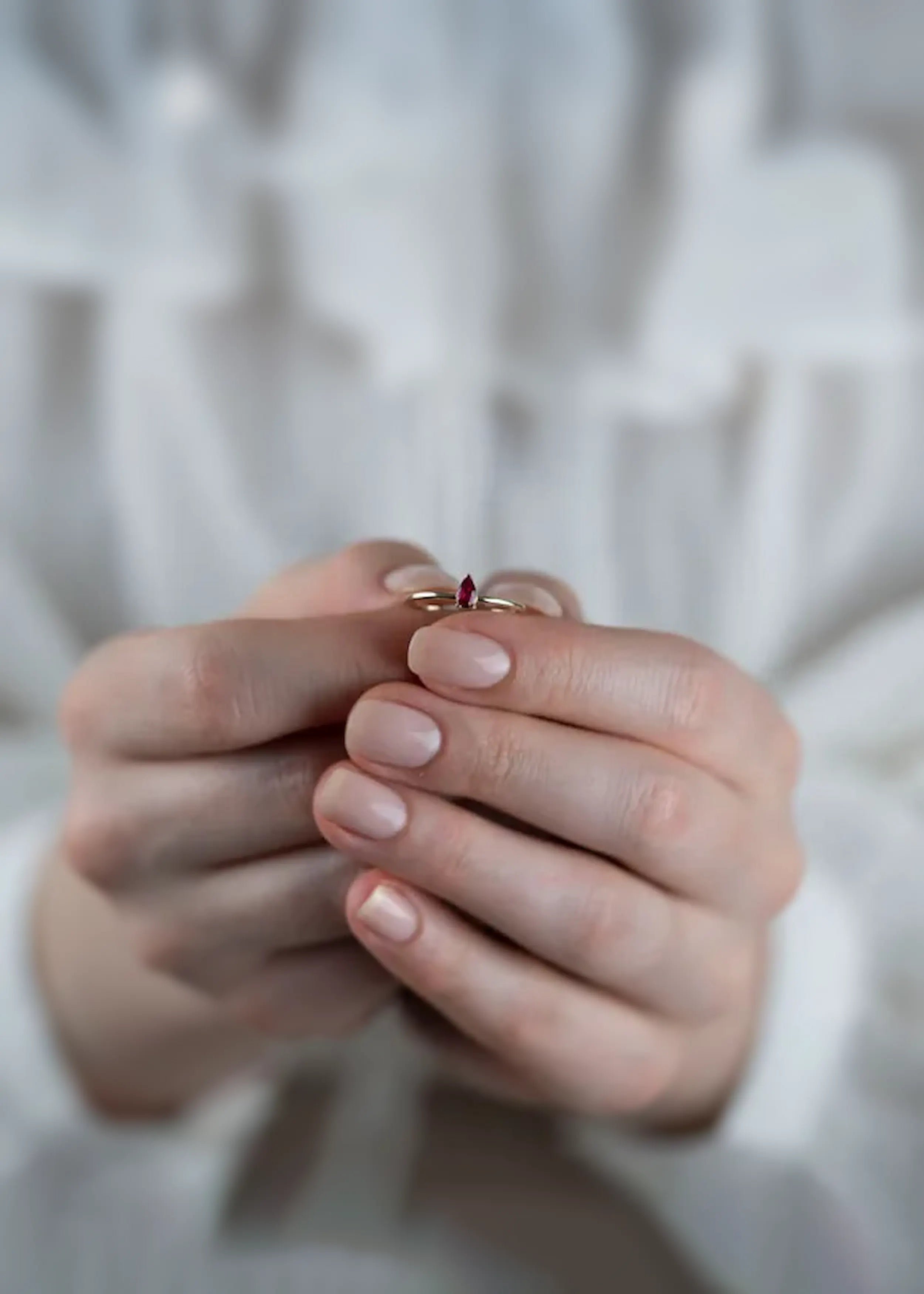 Goldener 14-Karat-Ring mit pear-förmigem pink-roten Stein, gehalten in den Händen einer Person mit naturfarbenen Nägeln. Die Person trägt ein weißes Kleidungsstück.