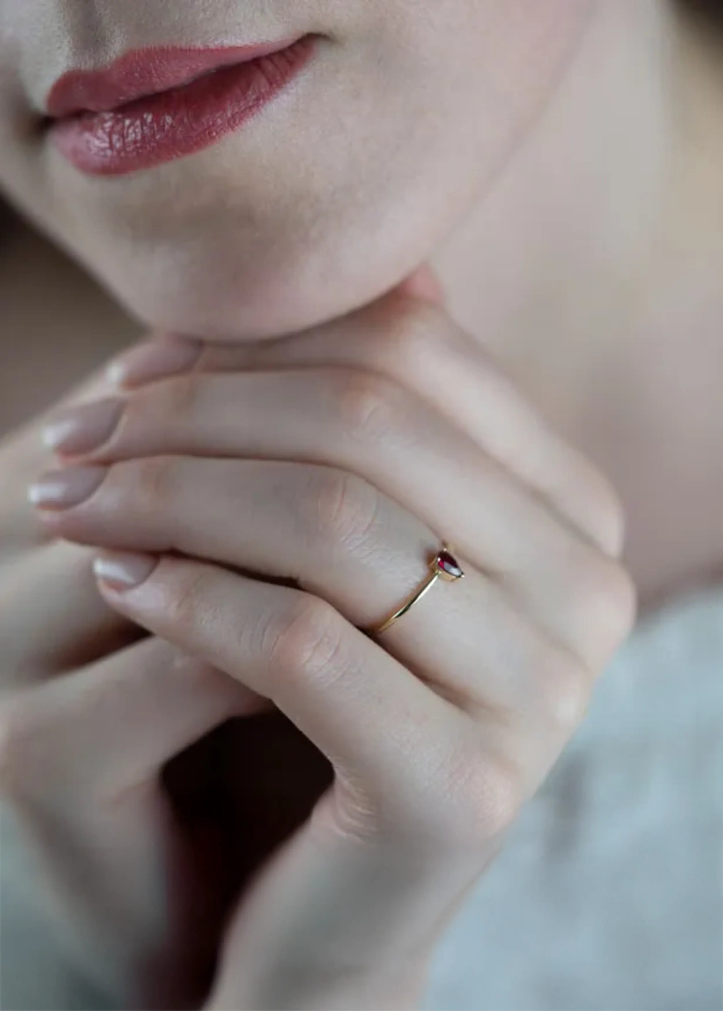 Goldener 14-Karat-Ring mit pear-förmigem pink-roten Stein, gehalten in den Händen einer Person mit naturfarbenen Nägeln. Die Person trägt ein weißes Kleidungsstück.