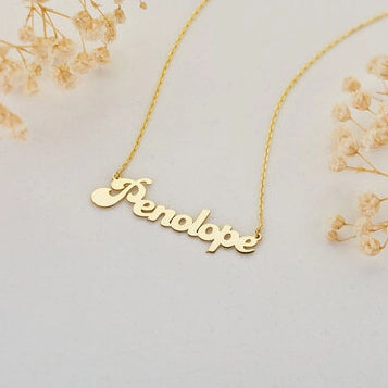 Filigrane Goldkette mit personalisiertem Namensanhänger 'PENELOPE' in eleganter Schreibschrift von Anadolu Juweliere, umgeben von getrockneten Blumen, die romantischen Schmuck in Düsseldorf hervorhebt