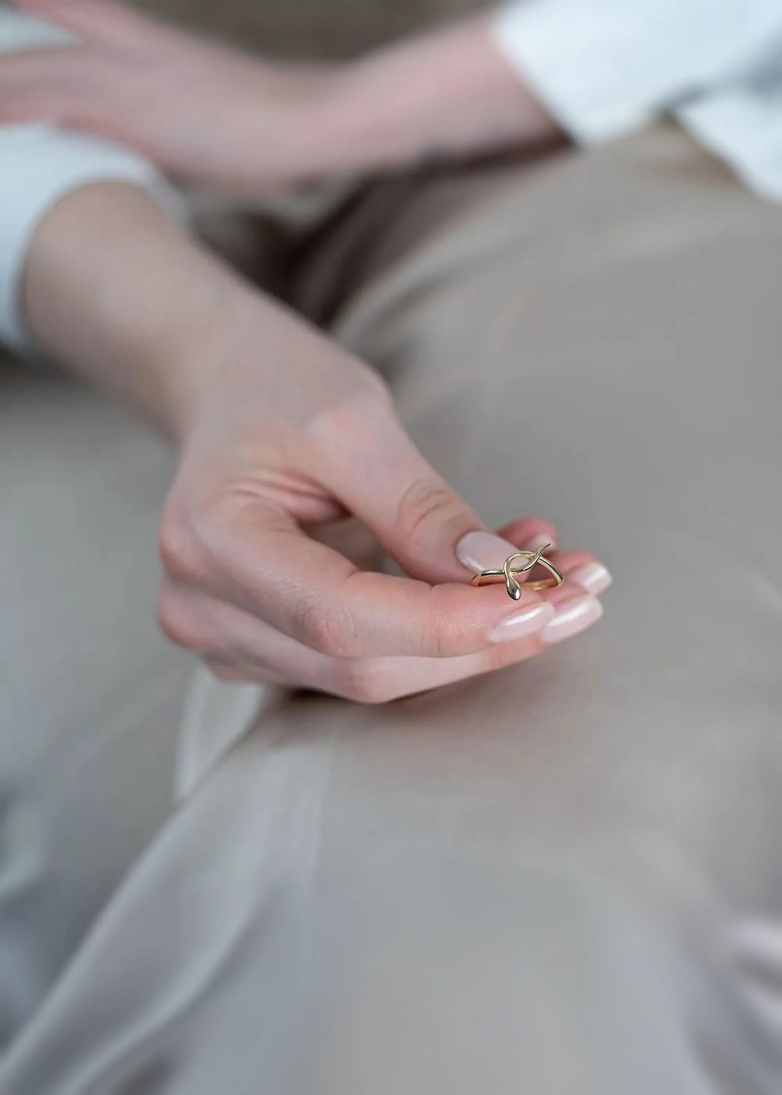 Goldener 14-Karat-Ring mit stilisiertem Schlangendesign, gehalten in der Hand einer Person mit naturfarbenen Nägeln. Die Person trägt ein weißes Kleidungsstück und sitzt auf beige Stoff.