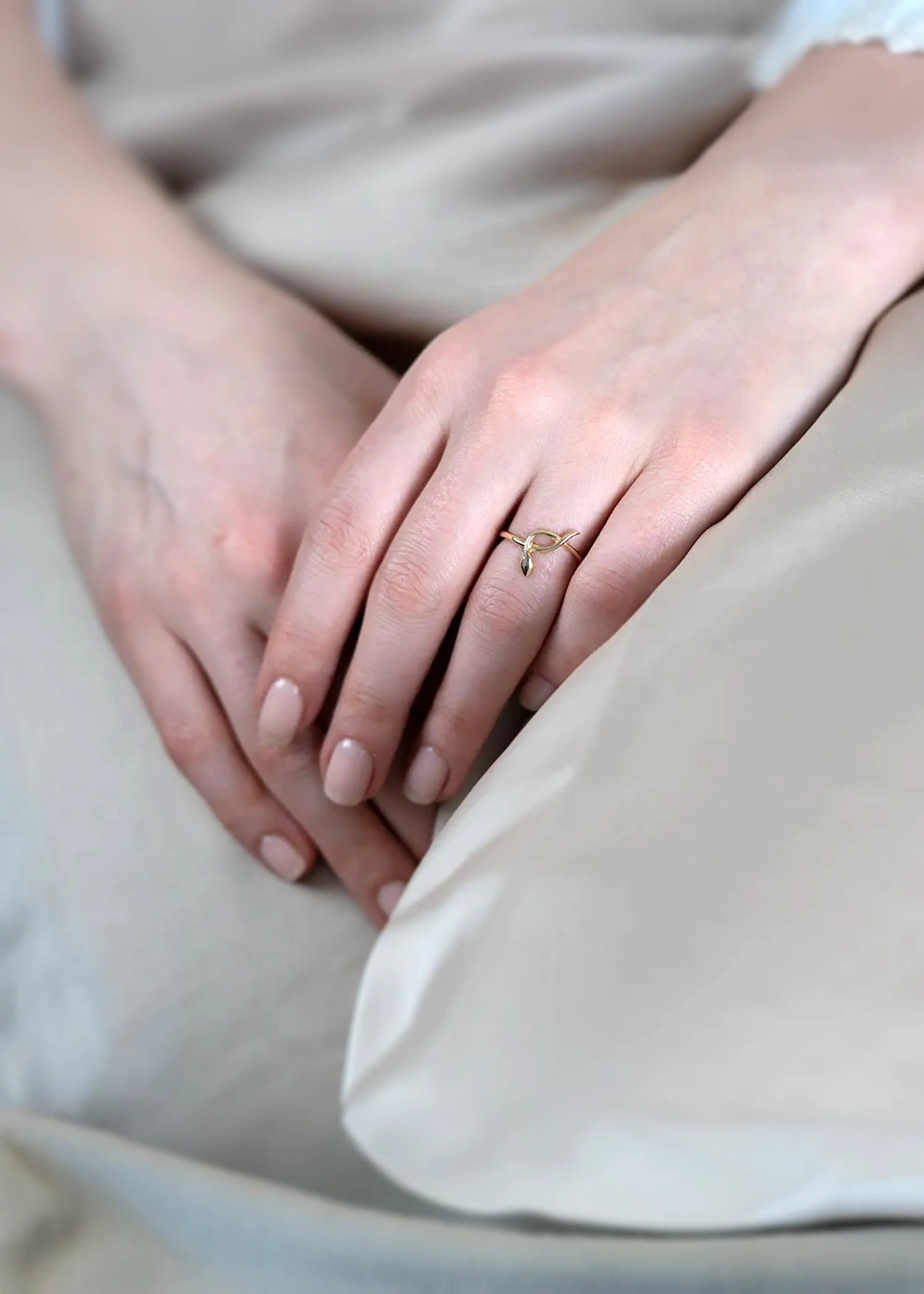 Goldener 14-Karat-Ring mit stilisiertem Schlangendesign, gehalten in der Hand einer Person mit naturfarbenen Nägeln. Die Person trägt ein weißes Kleidungsstück und sitzt auf beige Stoff.
