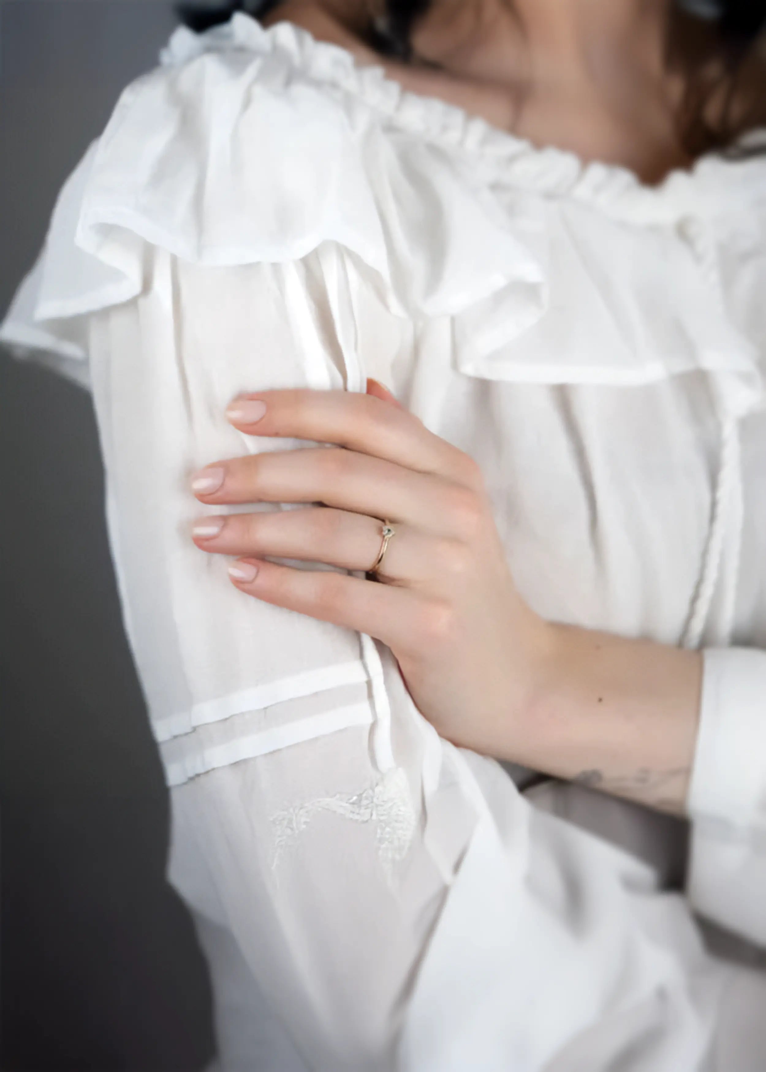 Goldener 14-Karat-Ring mit Blumenmotiv, einem grünen Stein im Zentrum und funkelnden Steinen, zwischen den Fingern einer Person gehalten. Die Person trägt ein weißes Kleidungsstück und hat naturfarbene Nägel, auf einem beige-weißen Stoff.