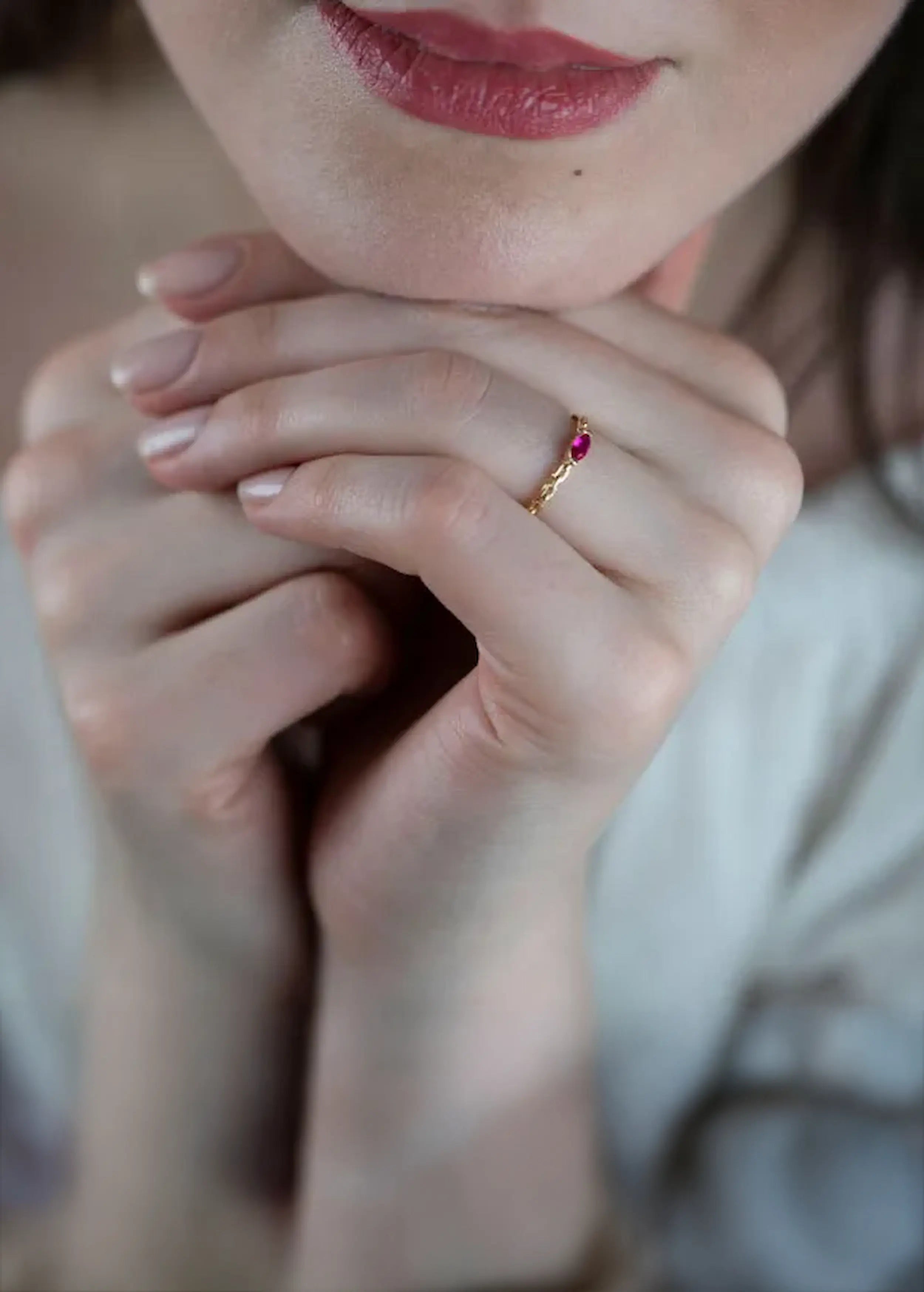Goldener Ring mit Gliederkette und einem ovalen pink-roten Stein, gehalten in den Händen einer Person mit naturfarbenen Nägeln. Die Person trägt ein weißes Kleidungsstück
